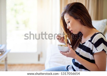 Young Asian woman sitting on her sofa at home enjoying a bowl of freshly made ramen noodles