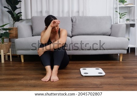 Young Asian woman sitting on the floor looking at the weight scale worried and depressed in dieting and eating disorder concept Imagine de stoc © 