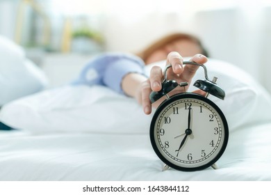 Young Asian Woman Sleeping On Bed Pressing Snooze Button On Black Vintage Alarm Clock At Seven O'clock Morning In Bed Room At Home, Lifestyle, Good Morning, Healthy Sleep And Joyful Weekend Concept