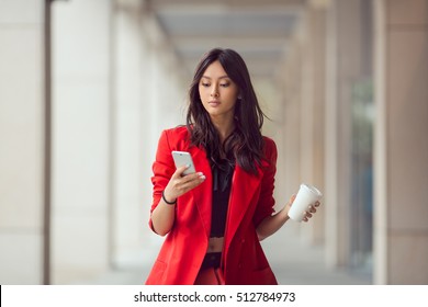 Young Asian woman with smartphone standing against street blurred building background. Fashion business photo of beautiful girl in red casual suite with phone and cup of coffee