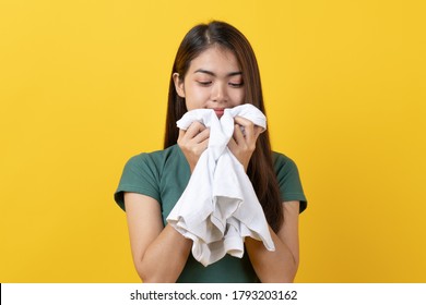 Young Asian Woman Smelling Scent Of Clean Clothes After Laundry Isolated On Yellow Background. Studio Shot.