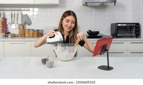 Young asian woman with a smile baking in a kitchen, using an electric mixer and a mixing bowl while looking at online cooking tutorial vdo on tablet computer. - Powered by Shutterstock