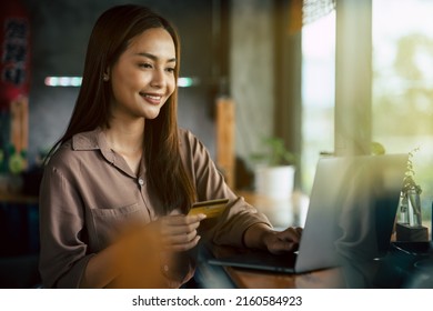 Young Asian Woman Smiling While Holding Credit Card In Her Hand For Doing Online Business With Laptop At Home. Relaxing Lifestyle Shopping  From Anywhere With Worry Free.