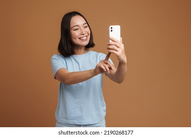 Young asian woman smiling while taking selfie on cellphone isolated over beige background