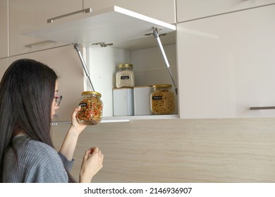 Young Asian Woman Stacks Glass Jars With Horns In Kitchen Cabinet. Storage Of Bulk Products. Zero Waste. Vegetarian Woman. Conscious Consumption.