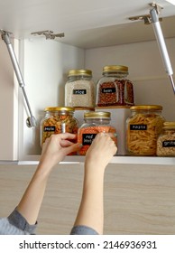 Young Asian Woman Stacks Glass Jars With Horns In Kitchen Cabinet. Storage Of Bulk Products. Zero Waste. Vegetarian Woman. Conscious Consumption.