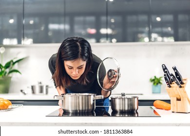 Young Asian Woman Standing Near Stove And Cooking.Happy Woman Looking And Smelling Tasting  Fresh Delicious From Soup In A Pot With Steam At White Interior Kitchen