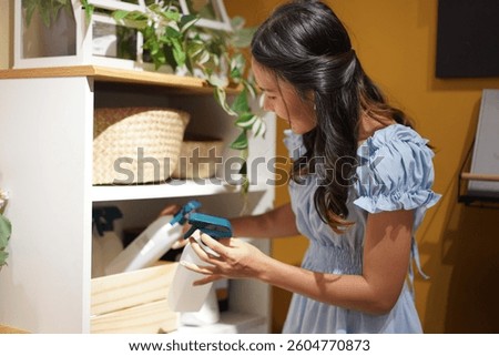 Young Asian woman standing near white shelf, organizing spray bottles and household items with concentration, casual home environment, wearing light blue dress, indoor lighting, tidy space