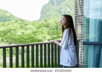 Young Asian woman standing on a balcony, enjoying the view of lush green mountains and nature. , looking relaxed and thoughtful in an outdoor setting with a scenic landscape in the background - Powered by Shutterstock