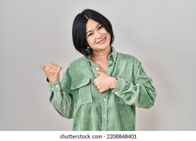 Young Asian Woman Standing Over White Background Pointing To The Back Behind With Hand And Thumbs Up, Smiling Confident 