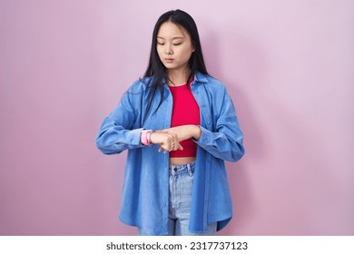 Young asian woman standing over pink background checking the time on wrist watch, relaxed and confident 