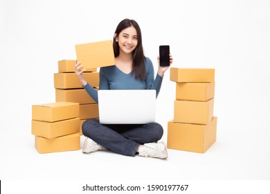 Young asian woman startup small business freelance holding parcel box, mobile phone and computer laptop and sitting on floor isolated on white background, Online marketing packing box delivery concept