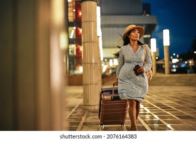 Young asian woman with straw hat and blue dress pulling suitcase in front of airport building
