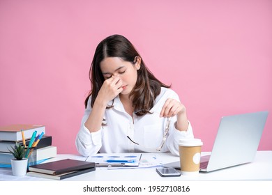Young Asian woman are stressed and tired from work. She was holding glasses at a white desk with a laptop in the office. Isolated on a pastel pink background.