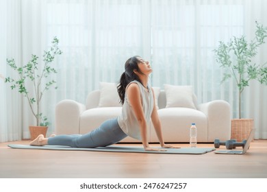 Young Asian woman stretching yoga workout on exercise mat while online training class with computer laptop in living room - Powered by Shutterstock