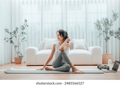 Young Asian woman stretching yoga workout on exercise mat while online training class with computer laptop in living room