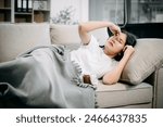Young Asian woman suffering from flu symptoms, covered with a blanket on a sofa. Concept of illness, healthcare, and recovery. Stay indoors, stay healthy

