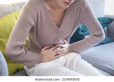 Young Asian woman suffering from strong abdominal pain, stomachache or period comes while sitting on sofa at home - Shutterstock ID 2154592635