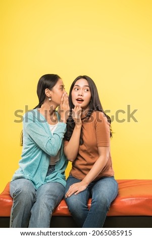 a young asian woman with a surprised gesture as her friend whispers while sitting together