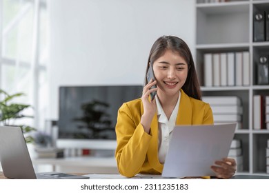 young asian woman talking on the mobile phone and smiling while sitting at her working place in office. - Shutterstock ID 2311235183