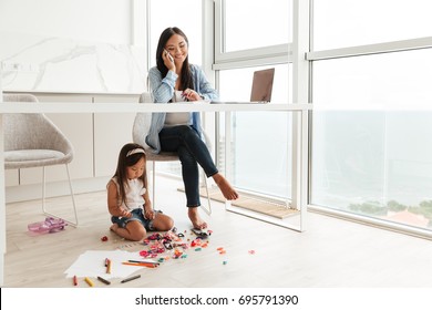 Young asian woman talking on a mobile phone and working on laptop while her little daughter playing on a floor at home