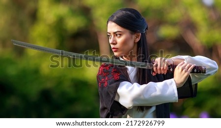 Young asian woman in traditional kimono trains in a fighting stance close-up portrait with katana sword samurai warrior girl in green summer garden 