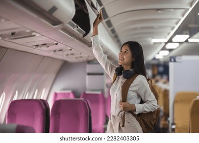 Young asian woman travel by airplane. Passenger putting hand baggage in lockers above seats of plane