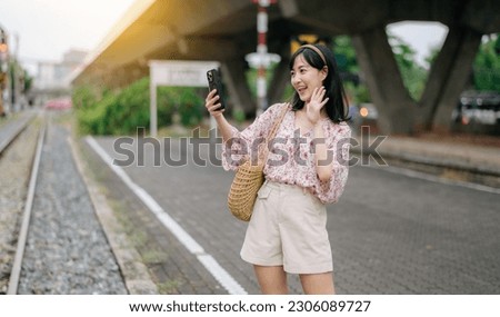 Young Asian woman traveler with weaving basket using a mobile phone beside railway train station in Bangkok. Journey trip lifestyle, world travel explorer or Asia summer tourism concept.