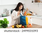 Young Asian woman unpacking groceries from blue shopper bag onto counter in kitchen