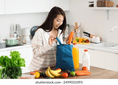 Young Asian woman unpacking groceries from blue shopper bag onto counter in kitchen - Powered by Shutterstock