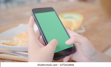 Young Asian Woman Using Black Mobile Phone Device With Green Screen. Asian Woman Holding Smartphone, Scrolling Pages While Sitting In Cafe. Chroma Key.