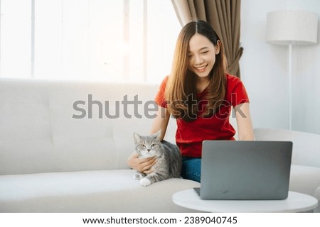 Young Asian woman using laptop sitting on couch at home, with relaxed sleeping ginger cat   
