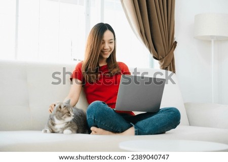 Young Asian woman using laptop sitting on couch at home, with relaxed sleeping ginger cat   