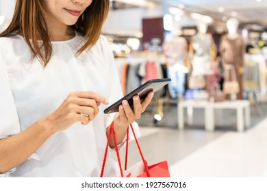 Young Asian Woman Using Mobile Phone While Holding Shopping Bag In Department Store. Consumerism And Lifestyle Concept