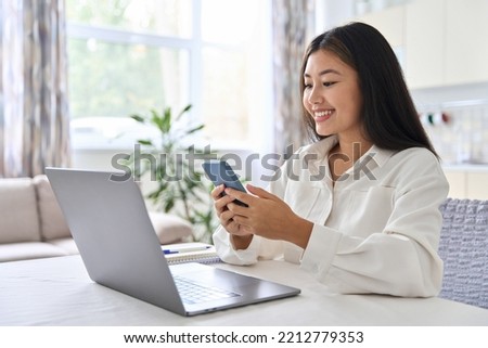 Young Asian woman using mobile phone while working at home with laptop. Female browsing site, messaging or shopping with smartphone during video call with friends on computer