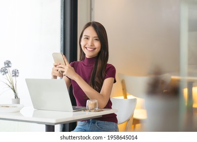 Young Asian Woman Using Phone And Laptop In Coffee Shop And Looking At Camera.