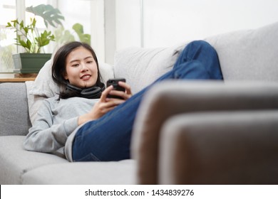 Young asian woman is using smartphone by connect with headset and lie down on the sofa. Relax, happy, technology concept