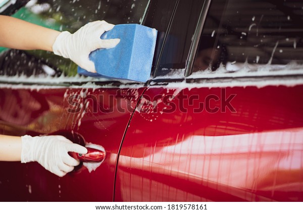 Young Asian woman using a soft sponge with a
liquid soap washing a car close up. Woman wash the modern red car
in the garage close up with copy
space.