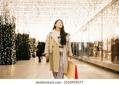 Young asian woman walking through a Christmas decorated shopping area, carrying shopping bags and enjoying the festive Christmas decorations. - Powered by Shutterstock