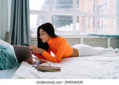 Young asian woman watching something on smartphone while lying and resting on bed with laptop and notebook at home. Domestic entertainment and leisure. Beautiful brunette girl of millennial generation