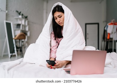 Young asian woman watching something on smartphone while sitting under blanket on bed with laptop. Domestic entertainment and leisure. Pretty brunette girl of millennial generation. Modern apartment