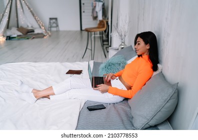 Young asian woman watching someyhing on laptop while lying and resting on bed at home. Domestic entertainment and leisure. Side view of brunette girl of millennial generation. Modern spacious flat