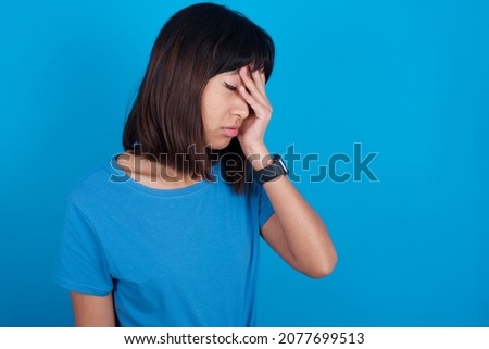 young asian woman wearing blue t-shirt against blue background with sad expression covering face with hands while crying. Depression concept.