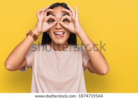 Young asian woman wearing casual clothes doing ok gesture like binoculars sticking tongue out, eyes looking through fingers. crazy expression. 