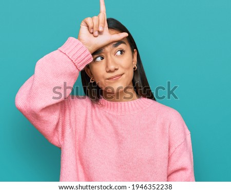 Young asian woman wearing casual winter sweater making fun of people with fingers on forehead doing loser gesture mocking and insulting. 