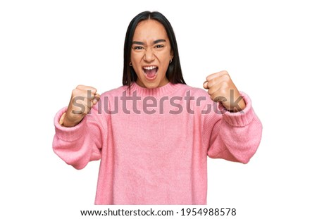 Young asian woman wearing casual winter sweater angry and mad raising fists frustrated and furious while shouting with anger. rage and aggressive concept. 