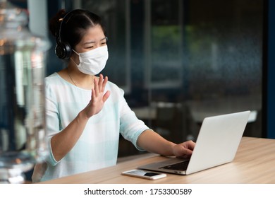 Young Asian Woman Wearing Face Mask And Headphones And Working Remotely By Using Computer Video Call Confernece At Coworking Space. Social Distancing And New Normal Lifestyle Concept