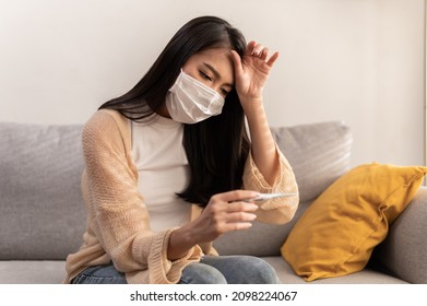Young Asian woman wearing face mask have a cold and high fever while checking body temperature by using digital thermometer. Daily lifestyle health care concept. - Shutterstock ID 2098224067