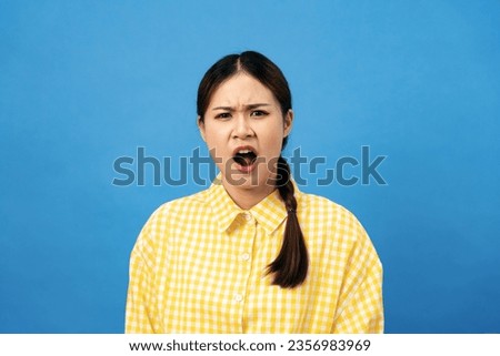 Young asian woman wearing gingham yellow shirt with braid hairstyle feeling angry and screaming isolated over light blue background. 