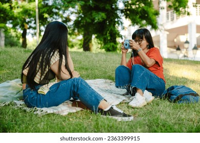 Young asian woman wearing glasses taking picture of her friend with instax camera while sitting on grass in green park